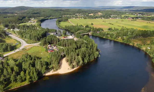 En vacker vy över en svensk landsbygd med en flod som slingrar sig genom gröna fält och skogar. En bro spänner över vattnet, och små röda hus syns bland träden. En idyllisk bild av sommar i Sverige.