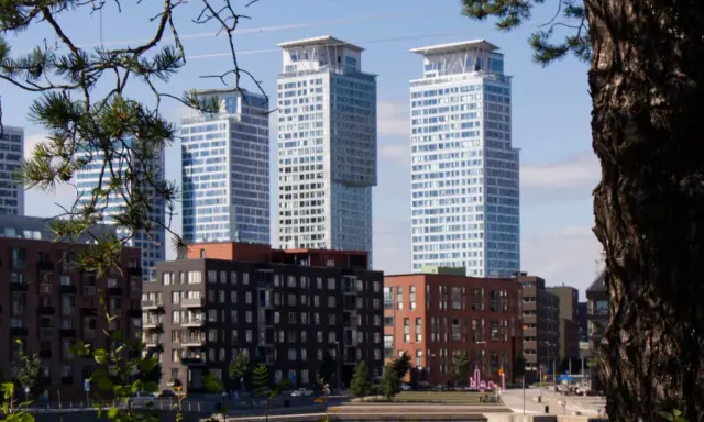 Höga, moderna skyskrapor reser sig bakom tegelbyggnader. En grönskande gren ramar in vyn, som om den undrar när sommaren startar i Sverige.