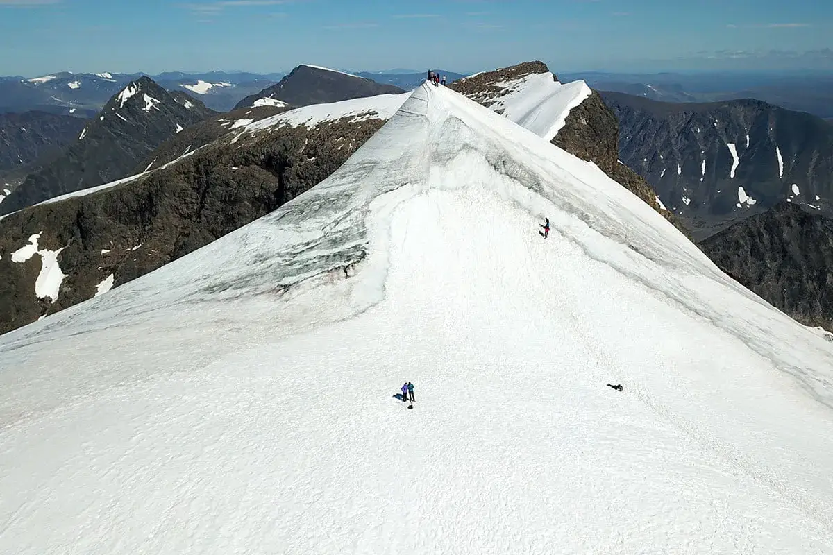 Vandrare på väg att bestiga Kebnekaise, en snöklädd bergstopp med dramatiska klippor och en vidsträckt utsikt.