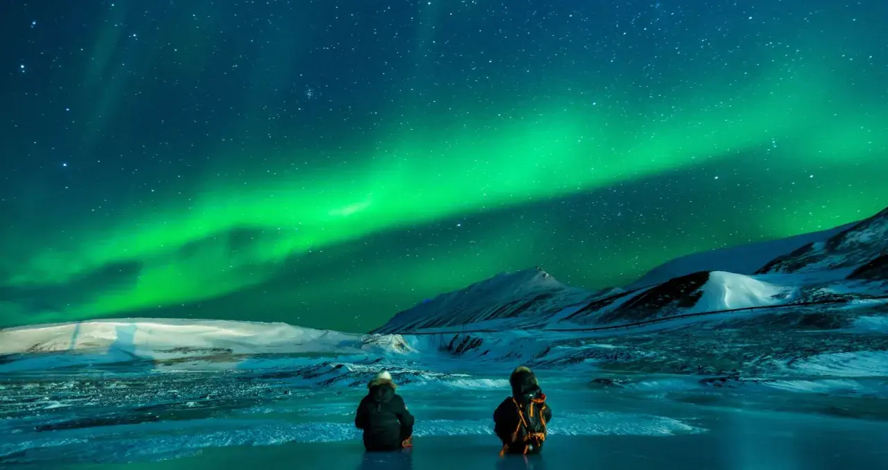 Två personer sitter på isen och ser på det magiska noorderlicht zweden. Snöklädda berg och stjärnor syns i bakgrunden.