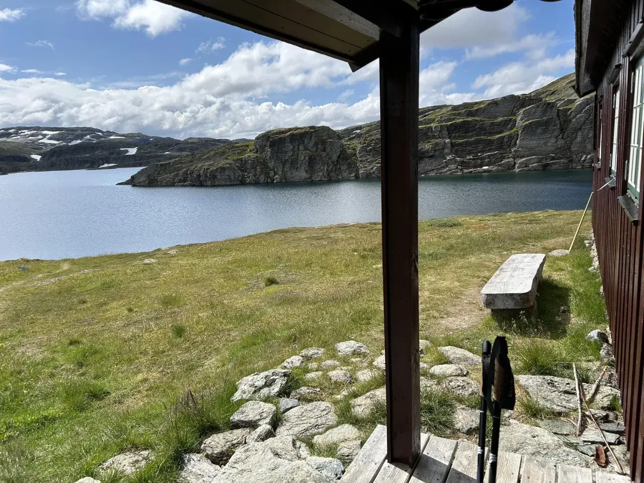 Vy från en stuga över en sjö och bergig terräng, perfekt för en Hardangervidda vandring. En bänk och vandringsstavar syns.