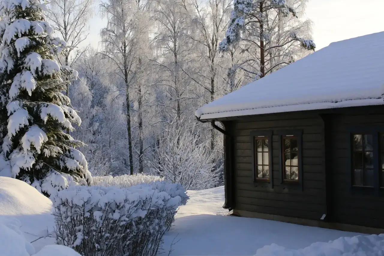 Vinterlandskap vid Kloten Nature Resort. Ett snötäckt hus med mörka träpaneler och fönster. Träd och buskar är täckta av frost och snö.