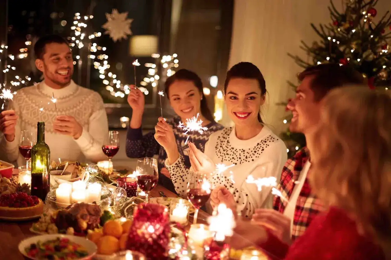 Vänner firar svensk jul med tomtebloss vid ett dukat bord med julmat och julgran i bakgrunden.