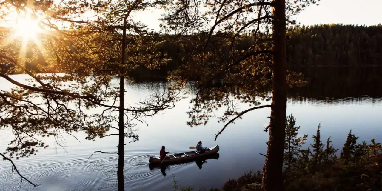 Två personer paddlar kanot på en lugn sjö i solnedgången, omgivna av skog. Ett stort leende sprider sig över himlen.
