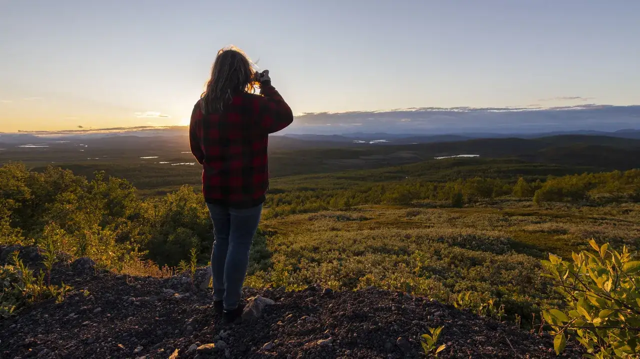 Kvinna med rutig skjorta fotograferar vid midnattssol i Sverige. Vacker vy över landskap med sjöar och skog i gyllene ljus.