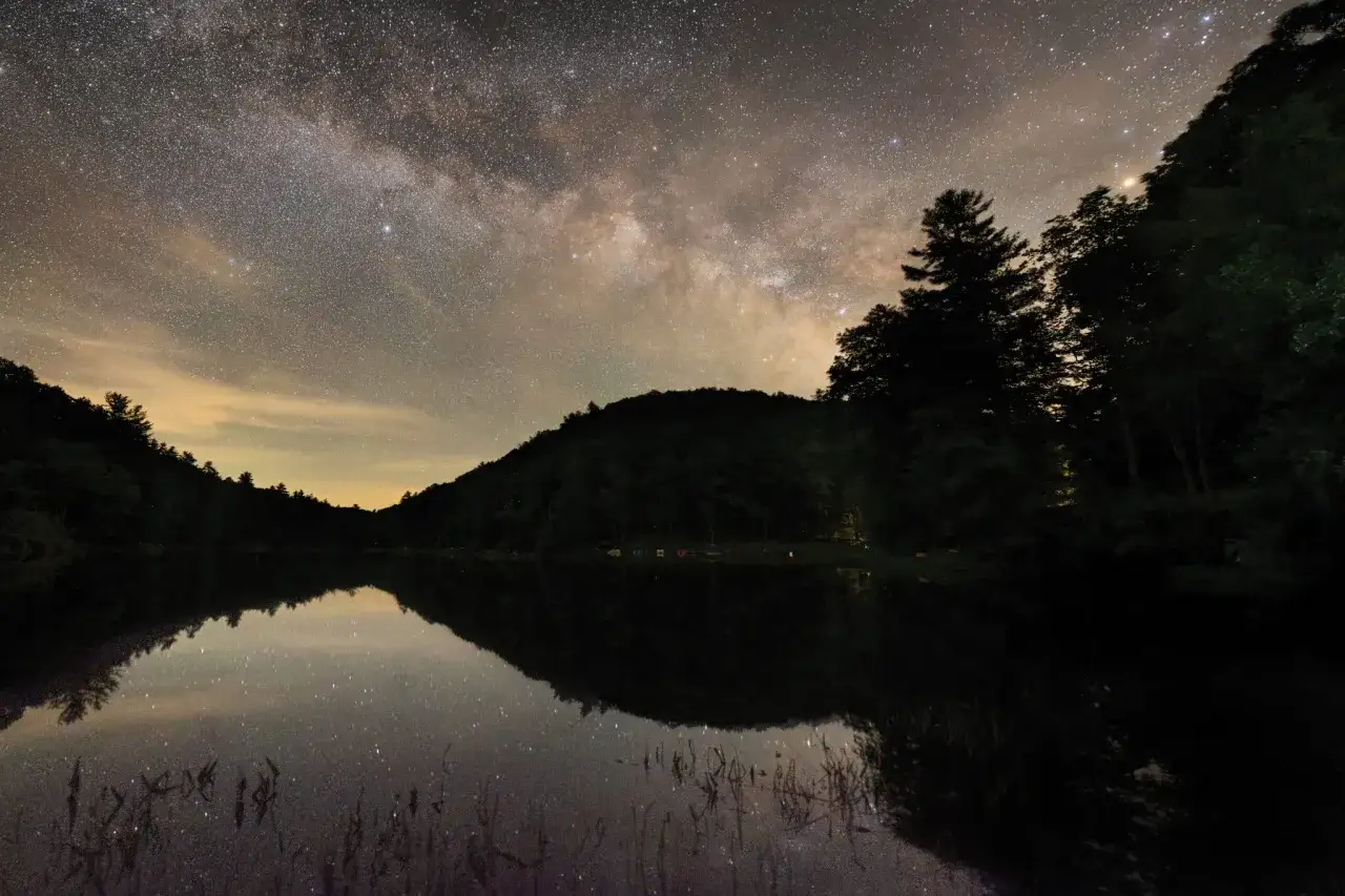 En spegelblank sjö reflekterar en stjärnklar natthimmel över skogsklädda kullar i Sverige.
