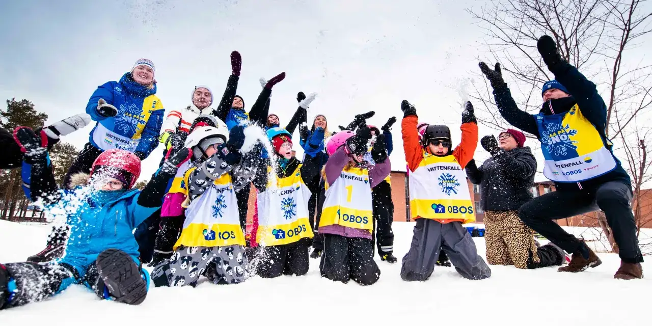 Glada barn och vuxna kastar snö i luften under sportlovsaktiviteter. Alla är klädda i varma kläder och västar med sponsortryck.