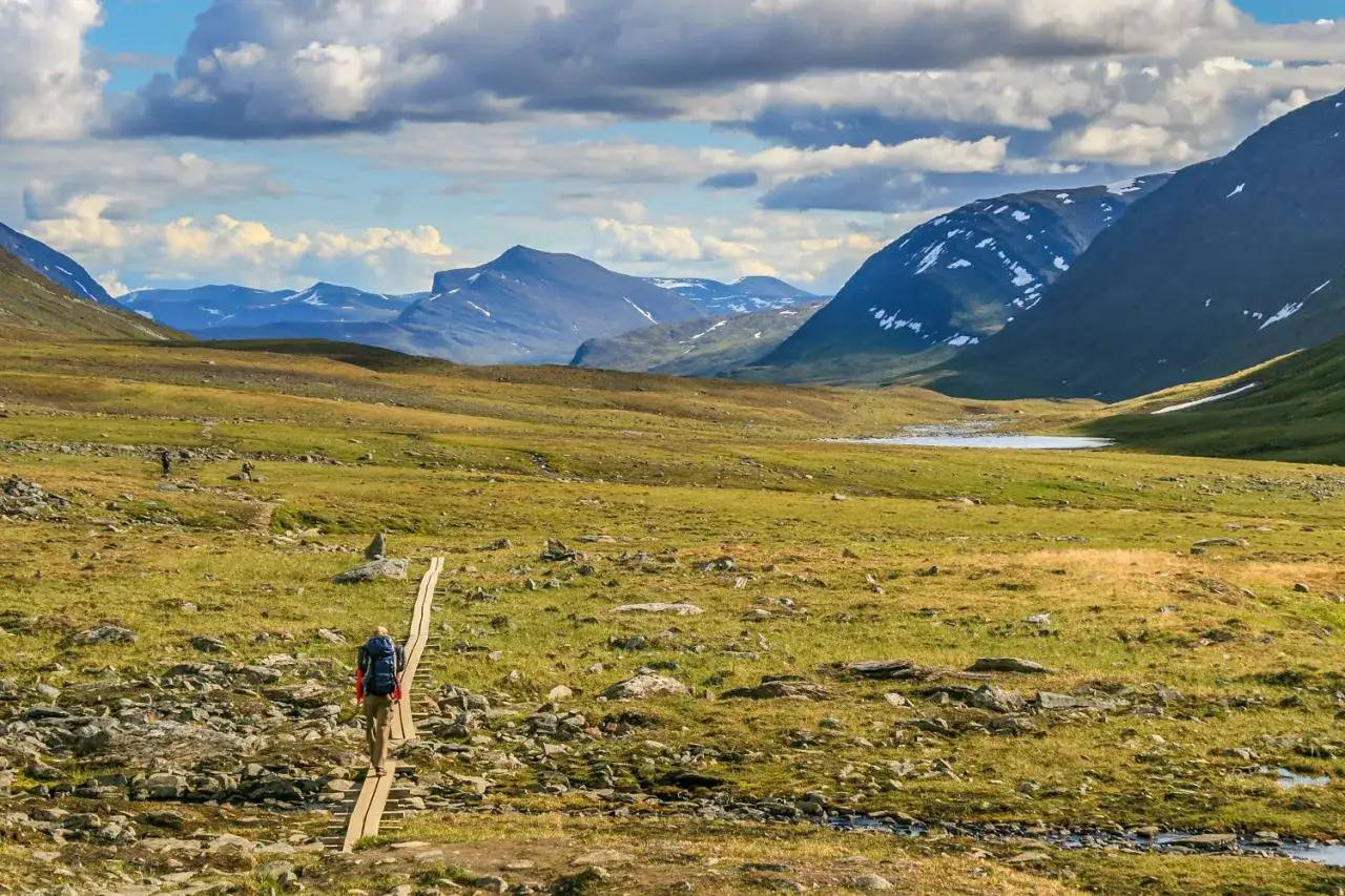 Vandrare på spång längs vägen från Nikkaluokta till Kebnekaise. Berg och moln i bakgrunden.