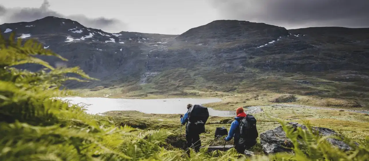 Två vandrare med stora ryggsäckar går ut på tur i ett dramatiskt fjällandskap med en sjö och snöfläckar.