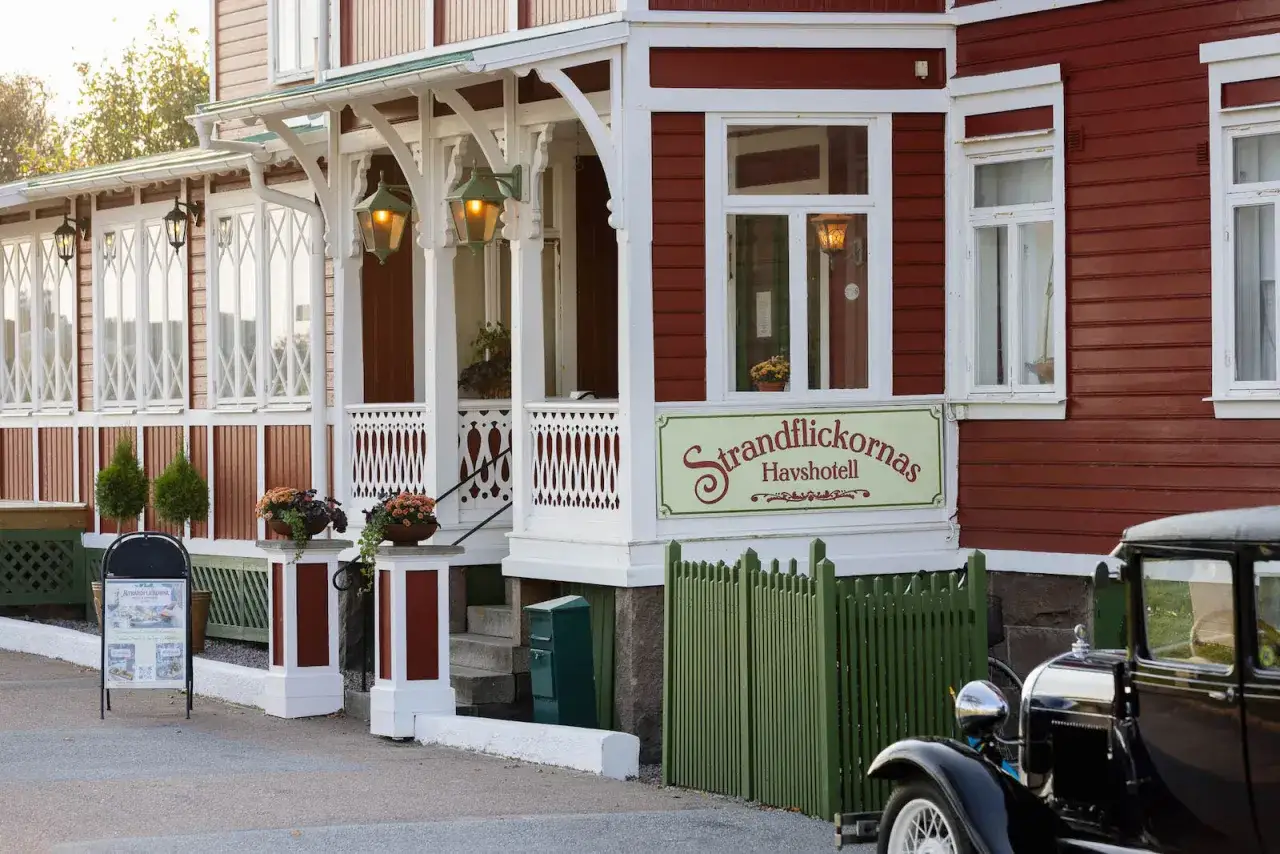 Vacker röd strandvilla med vita detaljer och en skylt för Strandflickornas Havshotell i Lysekil. En klassisk bil står parkerad utanför.