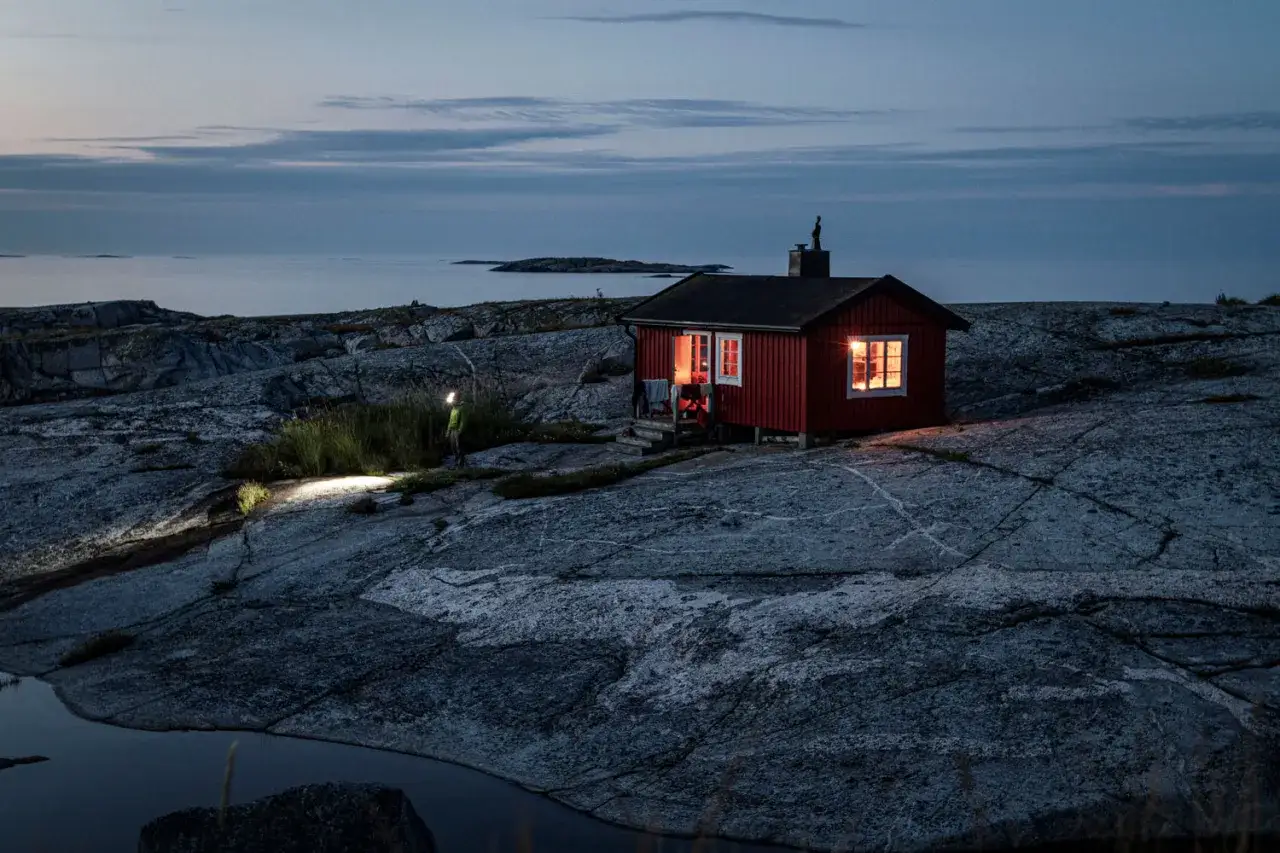 En person med pannlampa utforskar en klippig kustlinje vid skymningen. En röd stuga med upplysta fönster skapar en mysig känsla, perfekt för en utflykt Stockholm.