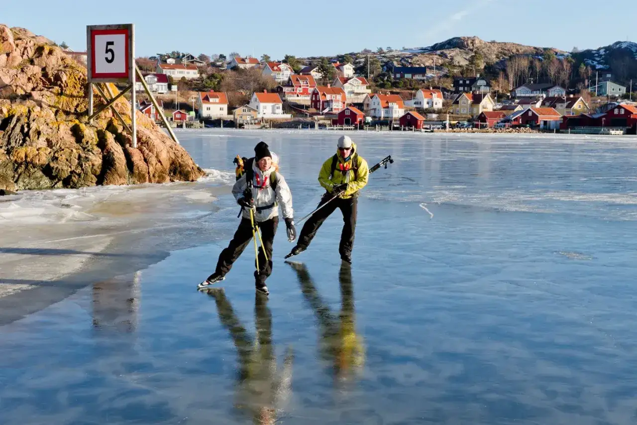 Två skridskoåkare på isen framför ett charmigt kustsamhälle. Frågan är hur tjock ska isen vara för att vara säker?