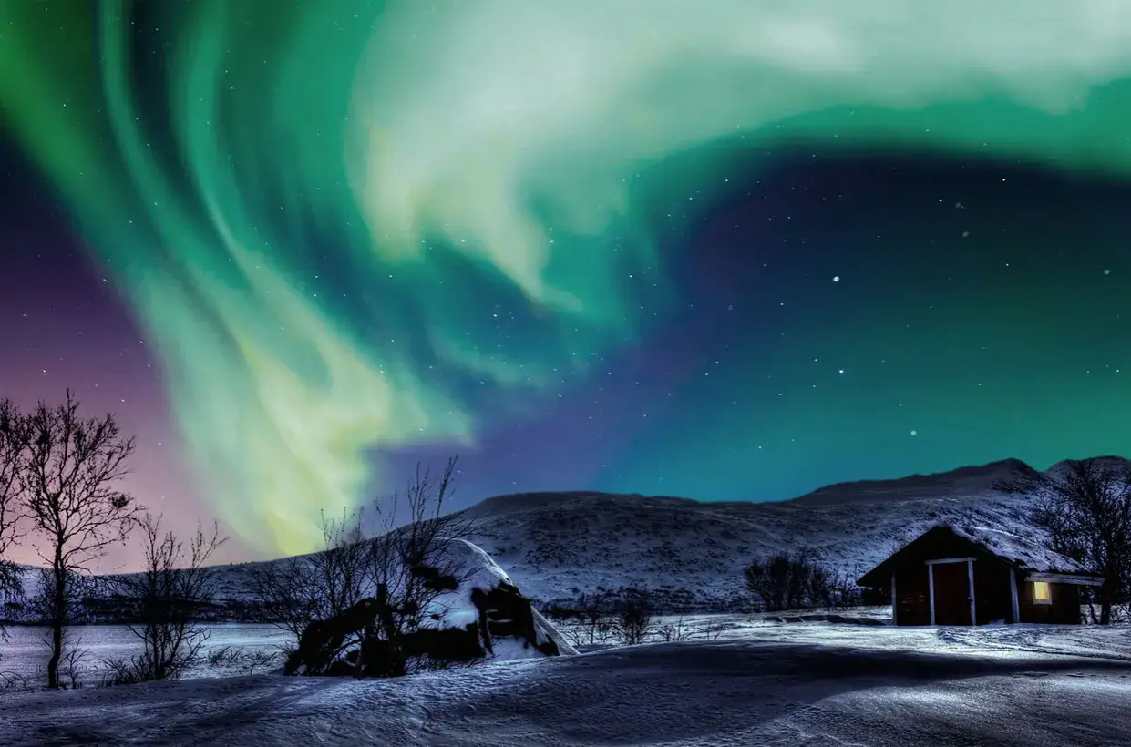 Norrsken dansar över snötäckt landskap med en liten stuga. En magisk vinternatt som visar en av Sveriges vackraste årstider.