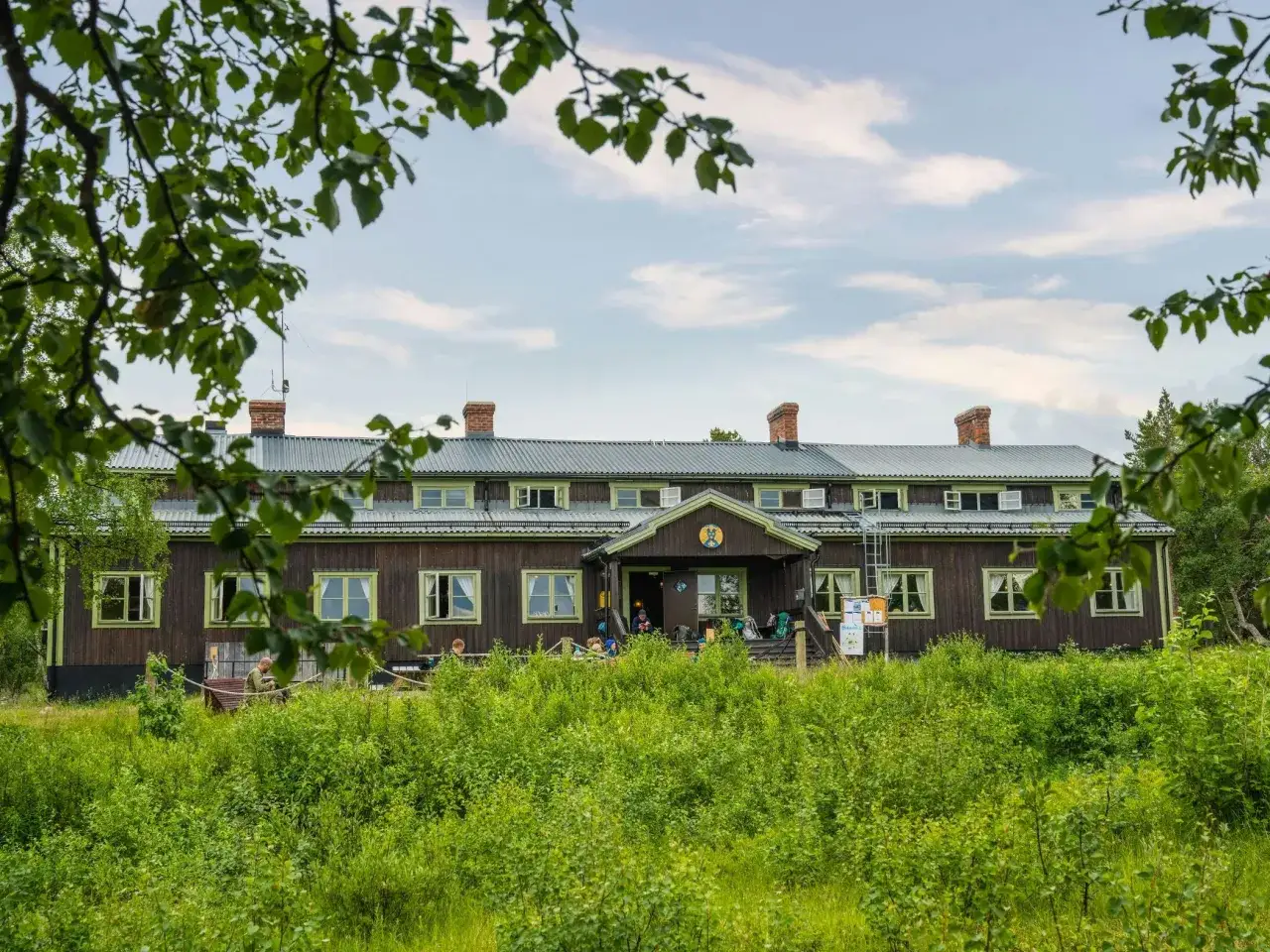 STF Saltoluokta fjällstation, en lång träbyggnad med gröna fönsterkarmar, omgiven av frodig grönska.