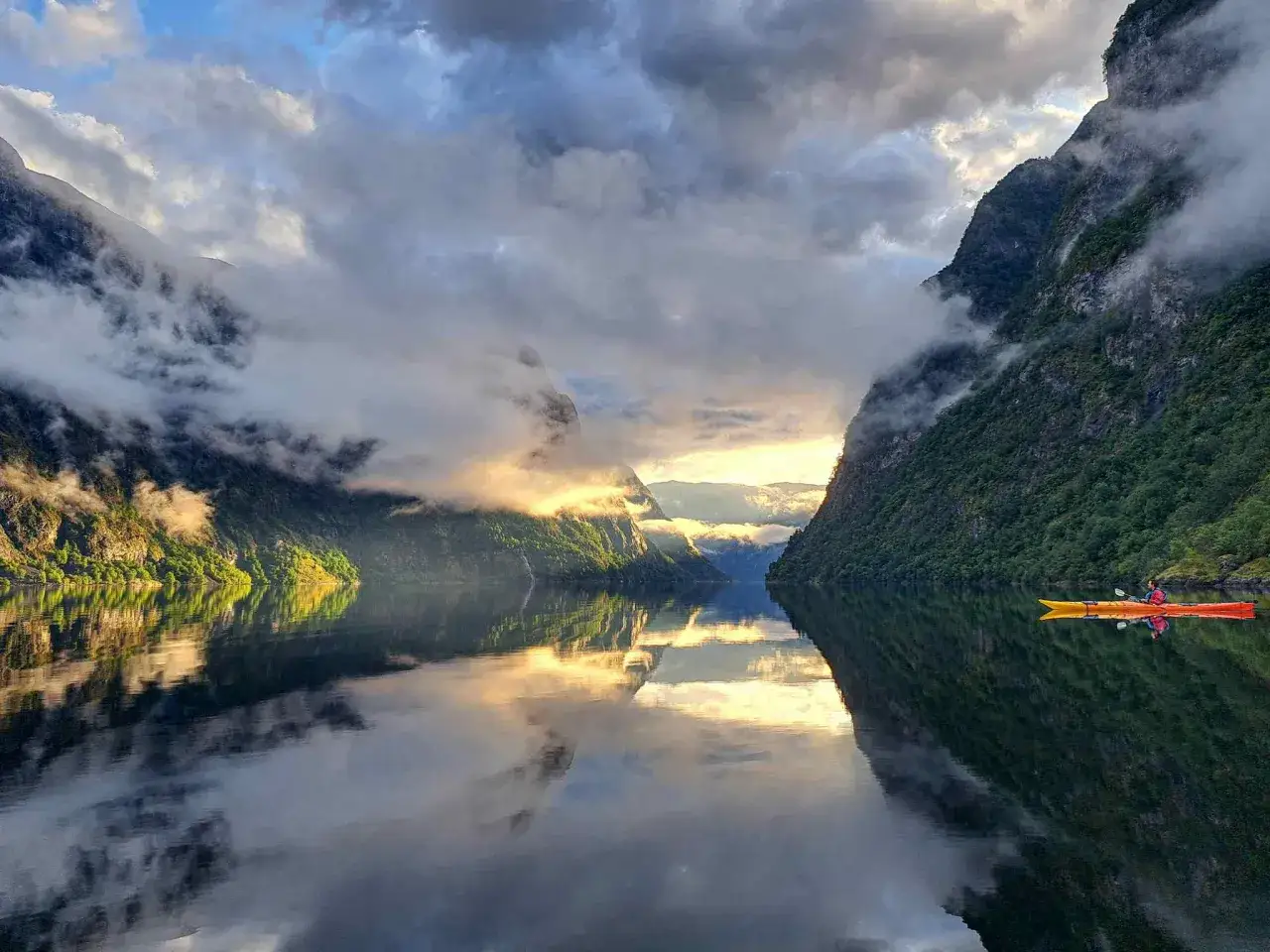 En kajakpaddlare njuter av den dramatiska natur Norge med moln som sveper över berg och speglar sig i det lugna vattnet.