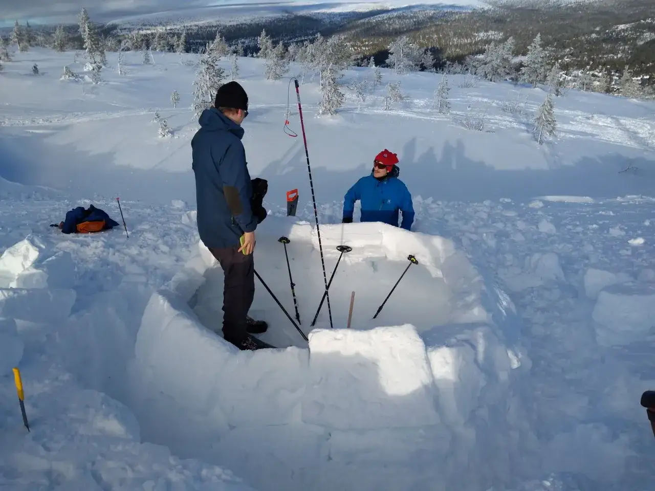 Två personer bygger en bivack i snön. De använder snöblock för att skapa väggar och en ingång. Skidor och stavar ligger i närheten.