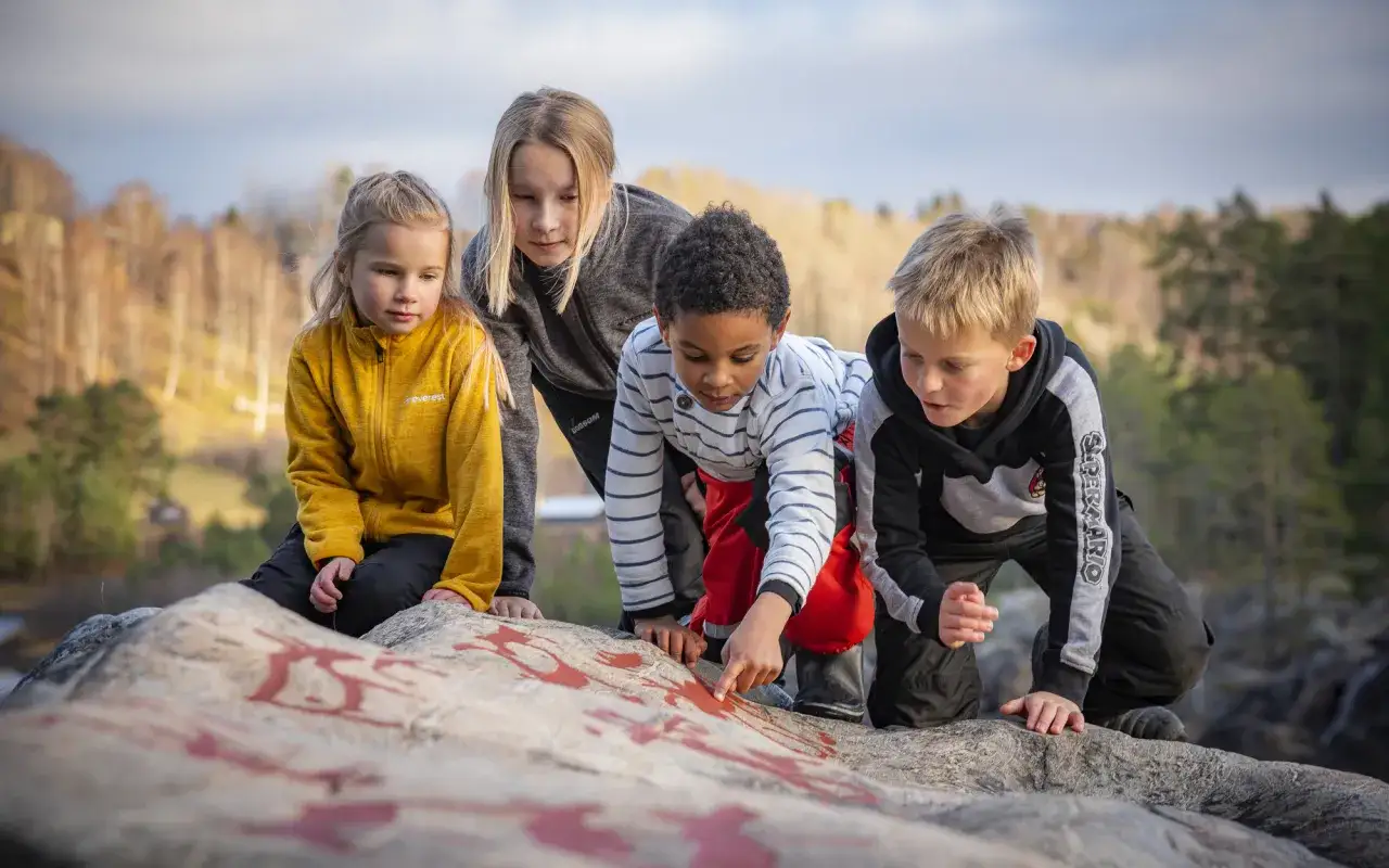 Fyra barn utforskar en sten med röda markeringar. En pojke pekar på stenens mönster. Utomhusaktiviteter för barn i naturen.