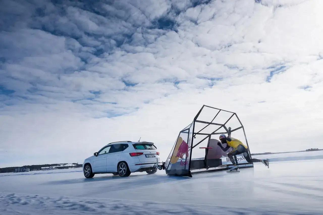 En skridskoåkare i gul tröja dras av en bil på isen vid Luleå. En glasbur med Red Bull-logga följer med.
