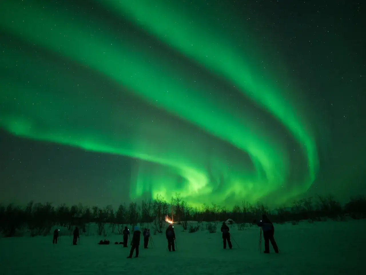 Människor ser norrsken i en snöig natt. En eld lyser upp bakgrunden, en påminnelse om att detta inte är det sydligaste midnattssol.