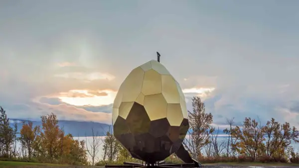 Solägg i Artipelag, en gyllene, fasetterad skulptur som liknar ett ägg, står mot en dramatisk himmel vid en sjö.