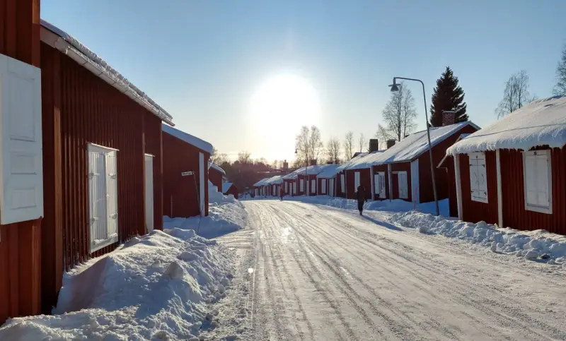 Vinterdag i kyrkbyn Gammelstad. Röda trähus med snö på taken kantar en snötäckt väg. Solen skiner starkt på en klarblå himmel.