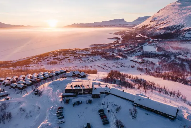 Vinterlandskap vid Kiruna med snötäckta fjäll, en frusen sjö och små stugor. Solen går ner och färgar himlen rosa.